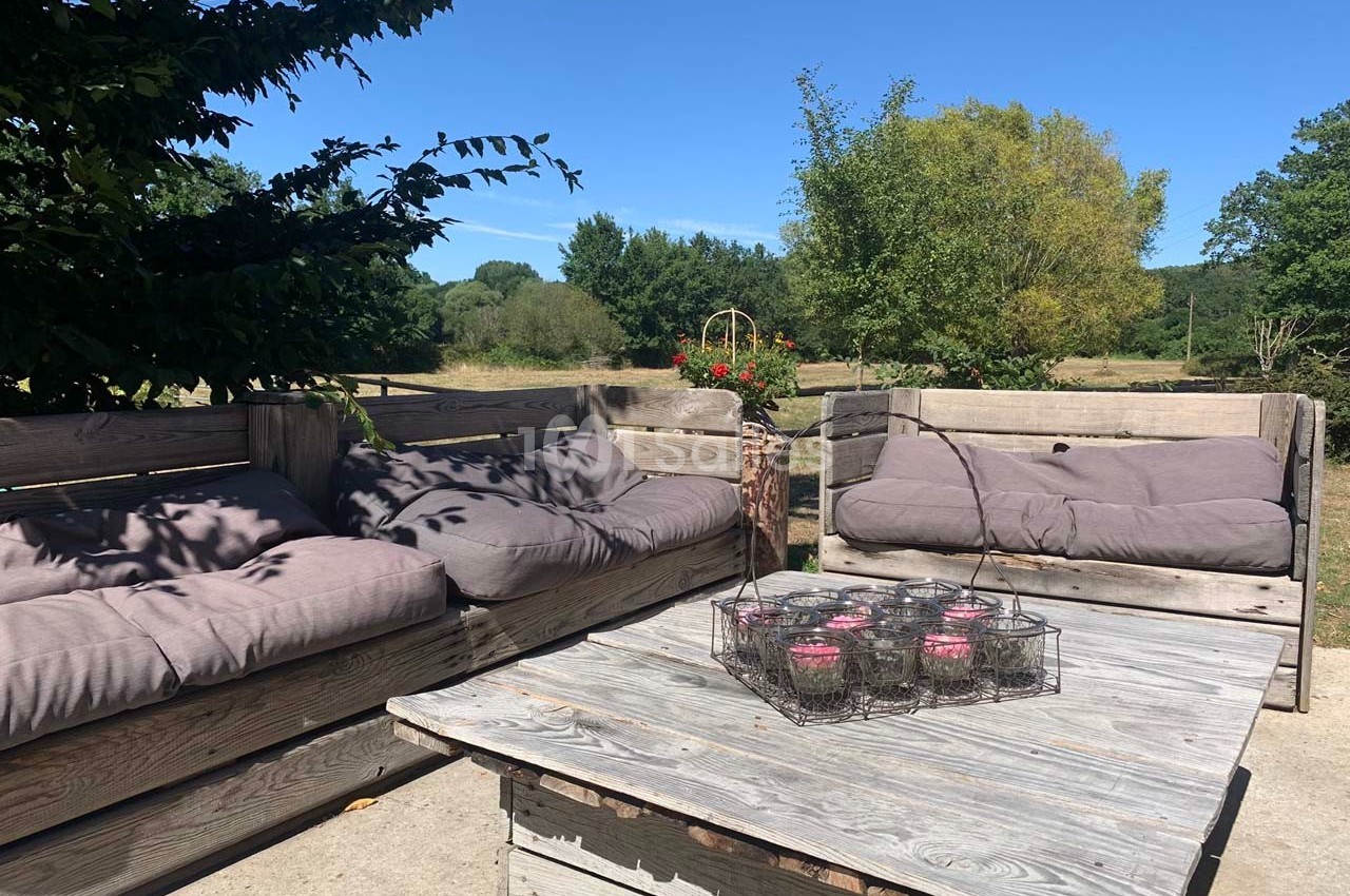Salon de jardin en bois avec coussins gris, table basse et bougies, situé en extérieur sous un ciel dégagé.