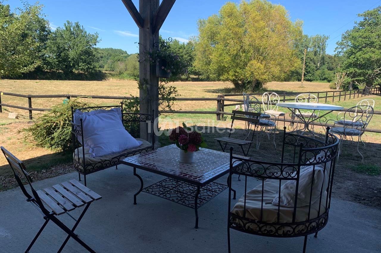 Salon de jardin en fer forgé sous une pergola, avec vue sur un champ clôturé et des arbres en arrière-plan.