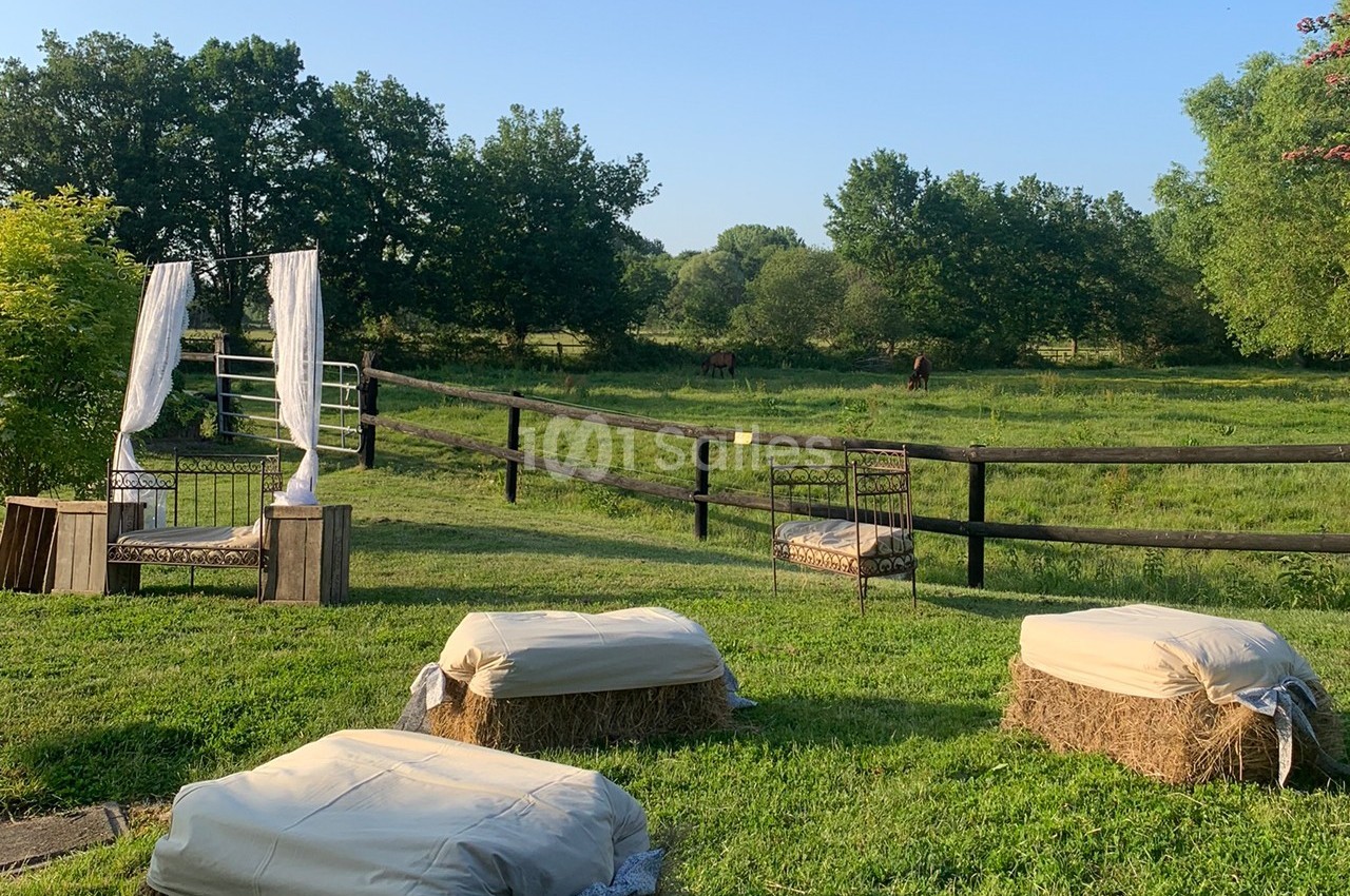 Bottes de foin recouvertes de tissu et fauteuils en fer forgé disposés dans un jardin avec vue sur un pré et des chevaux.