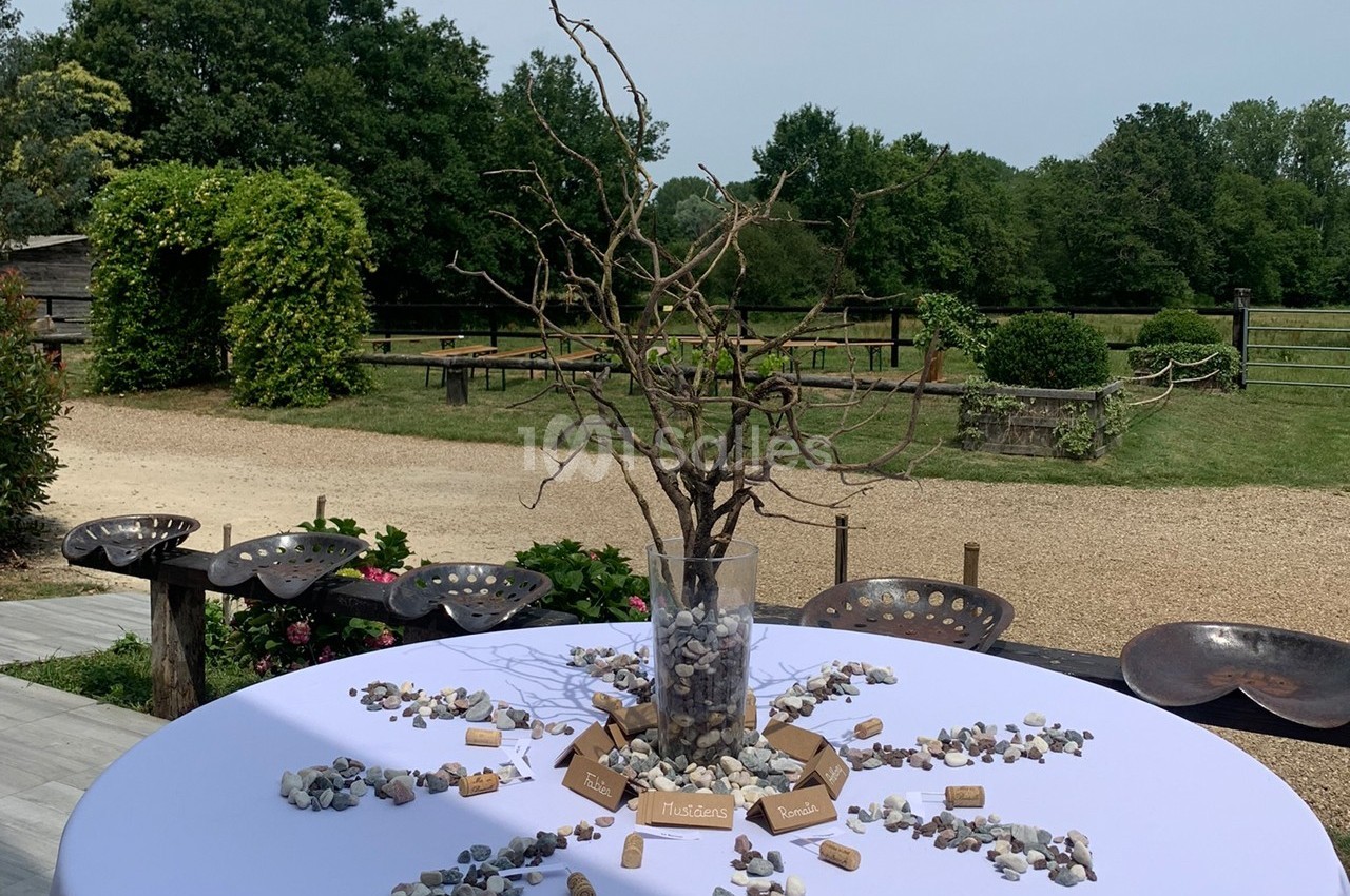 Centre de table avec branches sèches dans un vase, entouré de pierres et bouchons, sur une table ronde en extérieur.