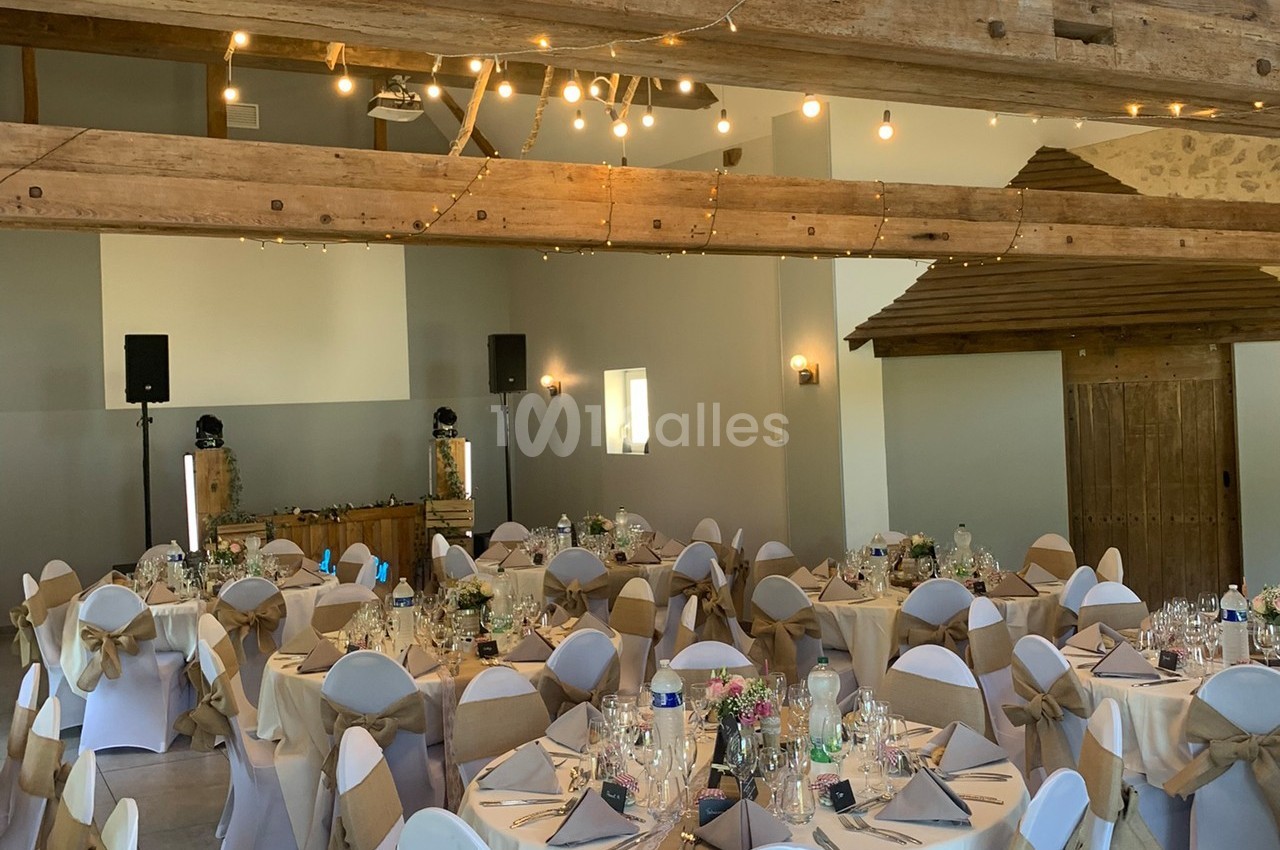 Salle de réception décorée avec des tables rondes dressées, des chaises ornées de nœuds et des guirlandes lumineuses…