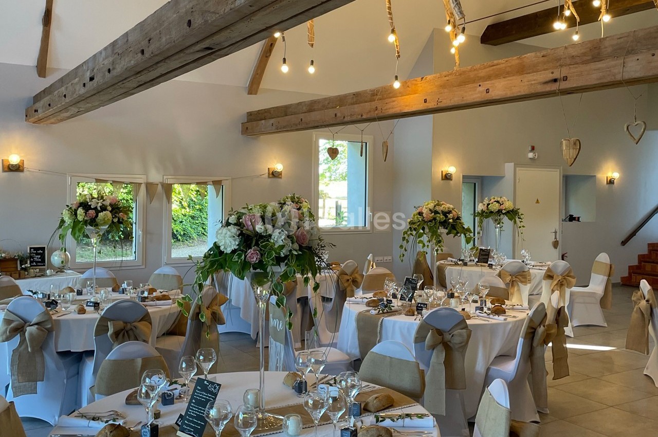 Salle de réception décorée pour un mariage, avec tables rondes, nappes blanches, fleurs et chaises ornées de rubans.