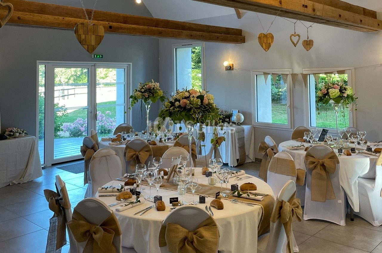 Salle de réception décorée pour un mariage, avec tables dressées, chaises ornées de nœuds et arrangements floraux.