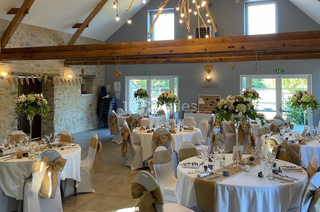 Salle de réception décorée pour un mariage, avec tables rondes, nappes blanches, chaises ornées de nœuds et compositions…