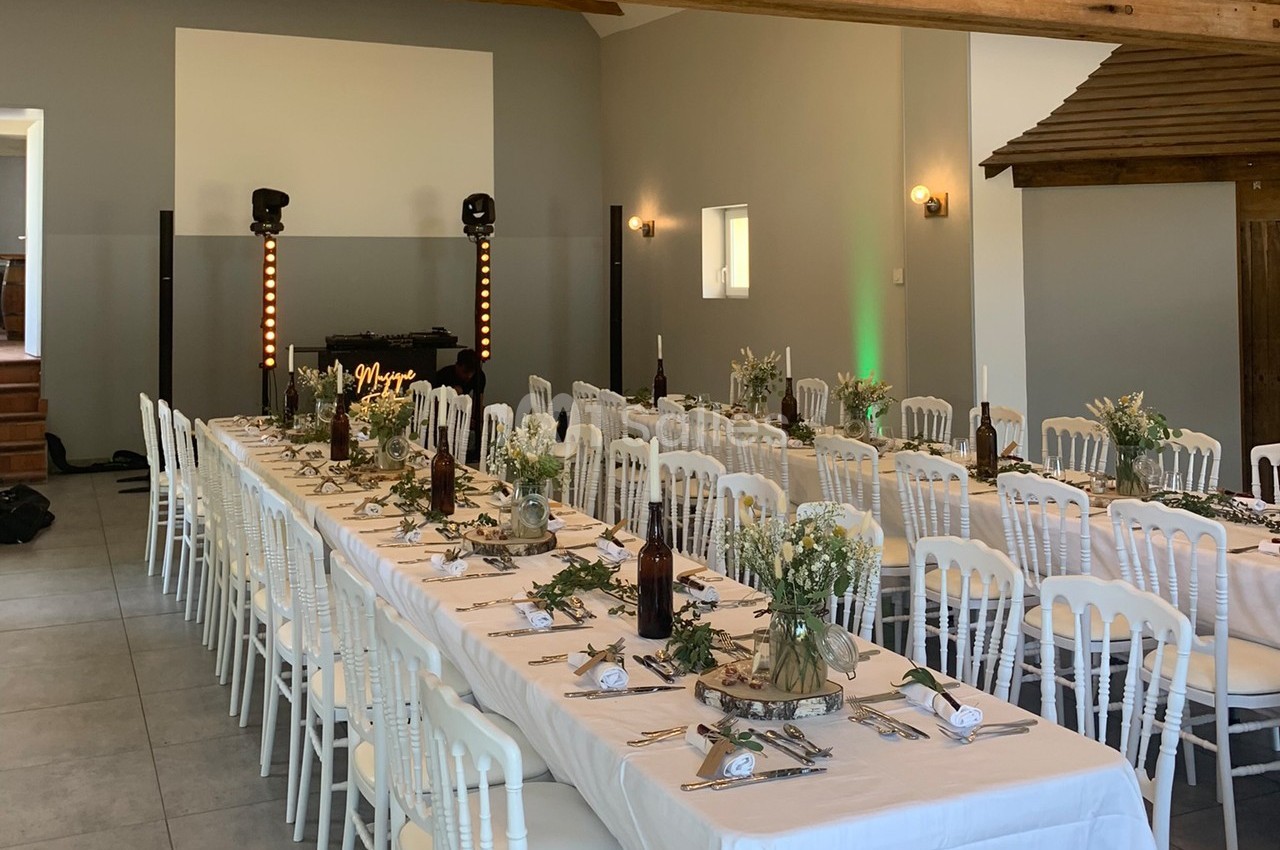 Salle décorée pour un repas festif avec longues tables dressées, chaises blanches et décoration florale.