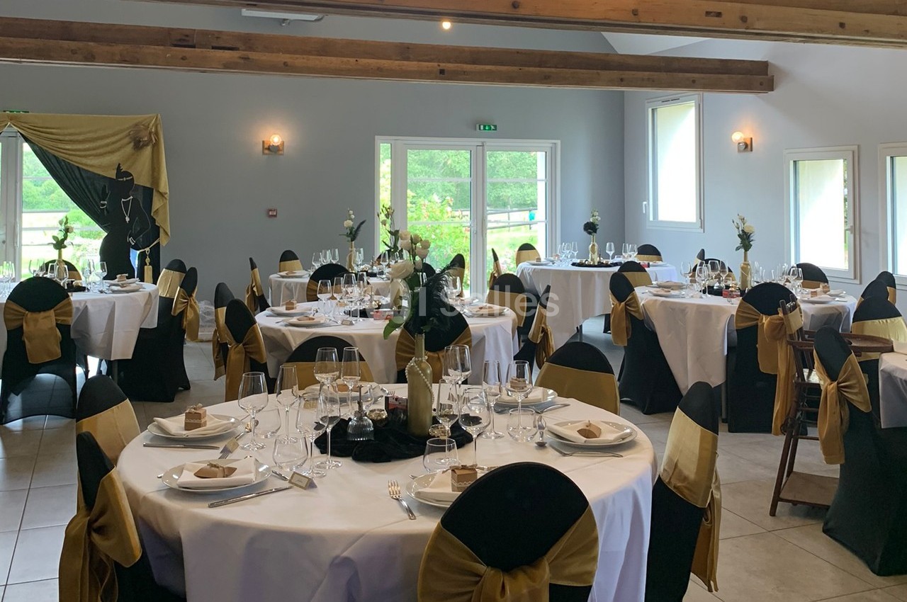 Salle de réception décorée avec des tables rondes, nappes blanches, chaises noires et dorées, et lumière naturelle.