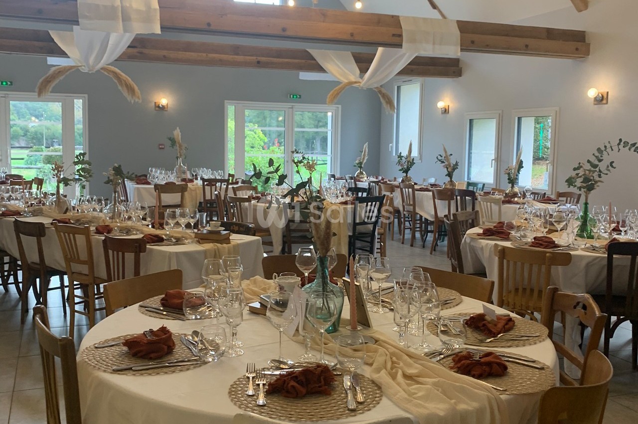 Salle de réception décorée avec des tables rondes dressées, nappes blanches, serviettes marron et éléments végétaux.