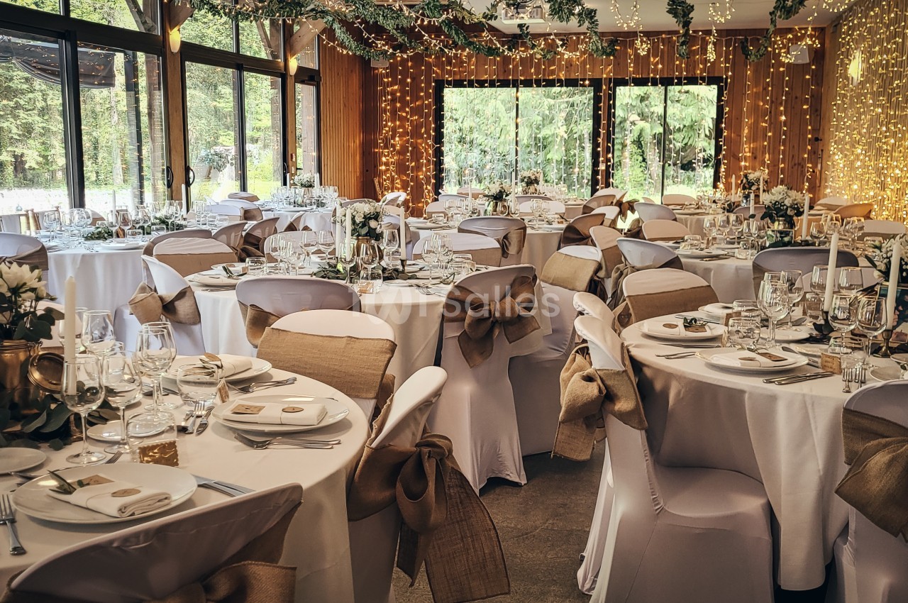 Salle de réception décorée pour un mariage, avec des tables rondes dressées, des chaises ornées et des guirlandes lumineuses.