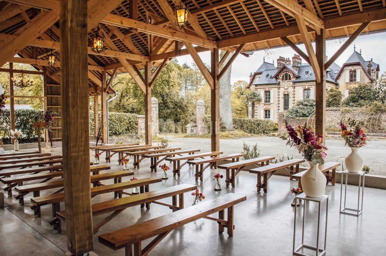 Structure en bois avec bancs alignés pour un événement, décorée de fleurs, avec un manoir en arrière-plan.