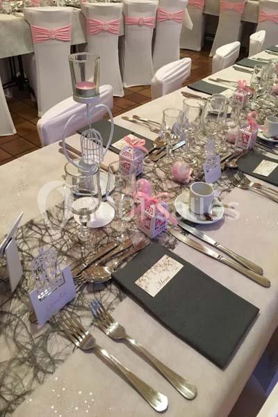 Table décorée pour un événement avec nappes blanches, chemins de table noirs, vaisselle élégante et décorations roses.