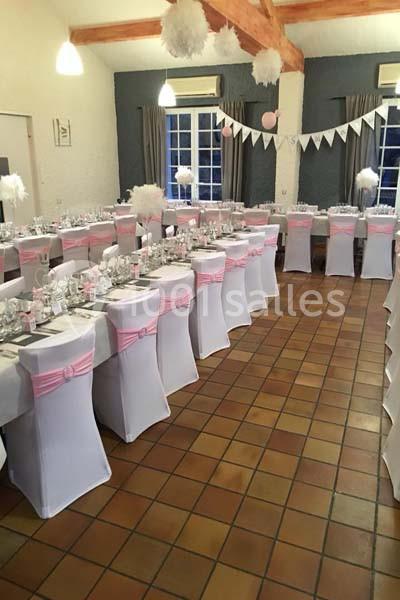 Salle décorée pour un événement, avec des chaises blanches ornées de nœuds roses et des tables dressées.