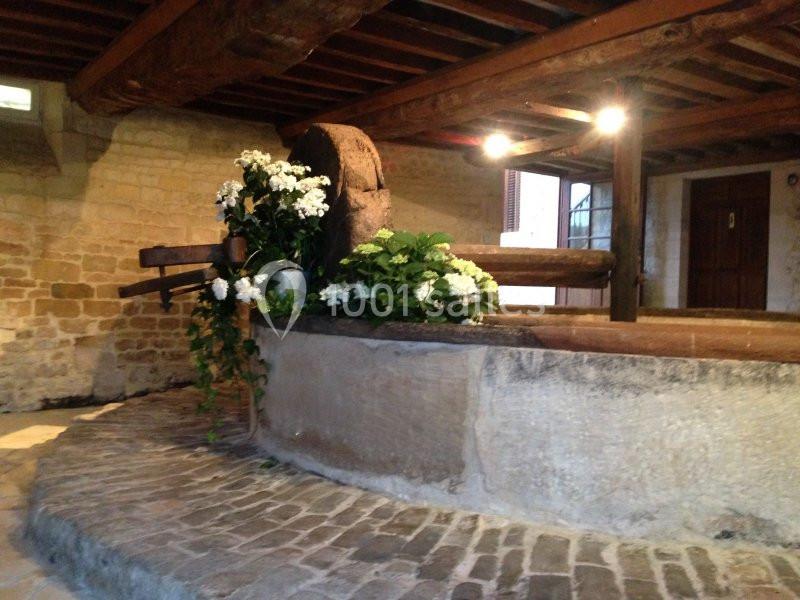 Ancien pressoir en bois dans une salle en pierre, décoré de fleurs blanches et entouré de pavés.