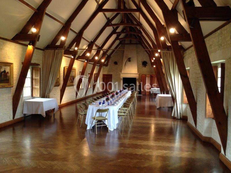 Grande salle avec une longue table dressée pour un repas, entourée de chaises, sous une charpente en bois apparente.