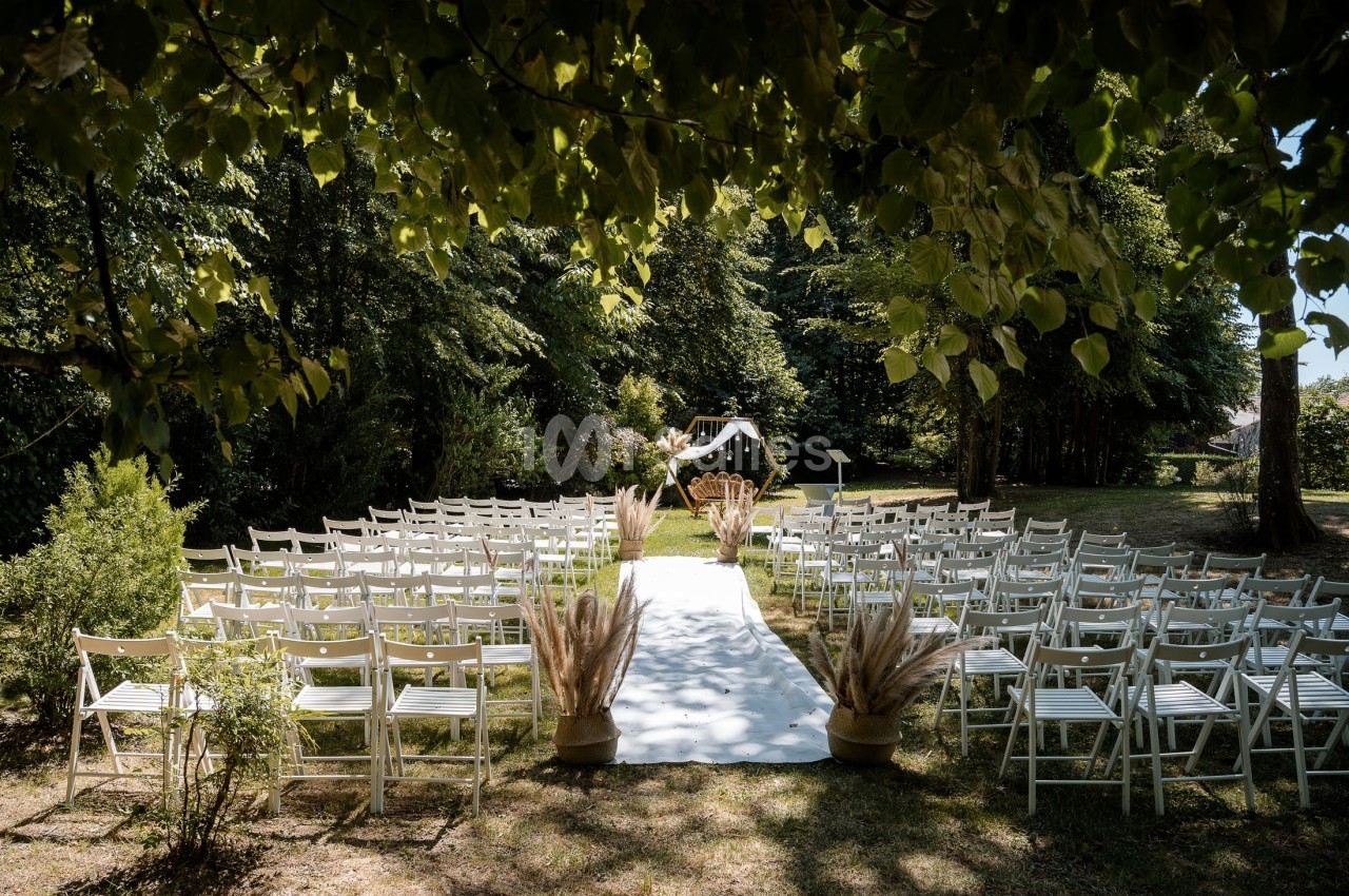 Allée centrale décorée pour une cérémonie de mariage en extérieur, entourée de chaises blanches et de verdure.