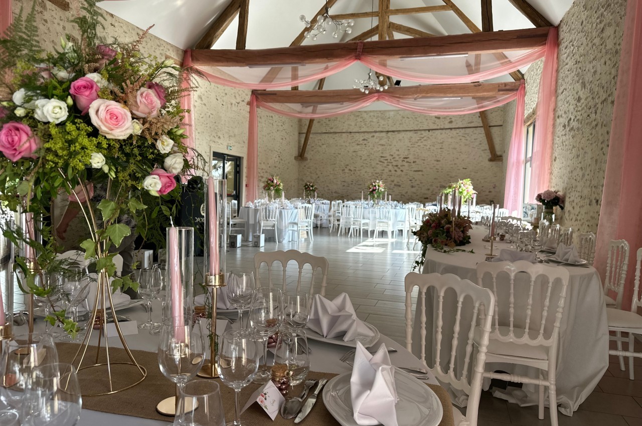 Salle de réception décorée avec des fleurs roses, nappes blanches et chaises, sous une charpente en bois apparente.