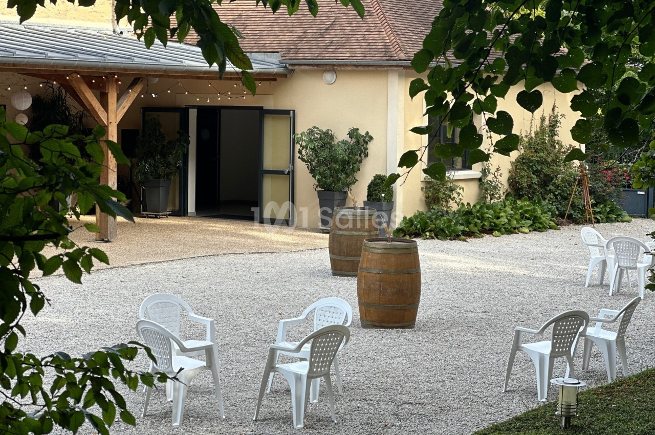 Cour extérieure avec gravier, chaises blanches en plastique, tonneaux en bois et végétation autour d'un bâtiment jaune.