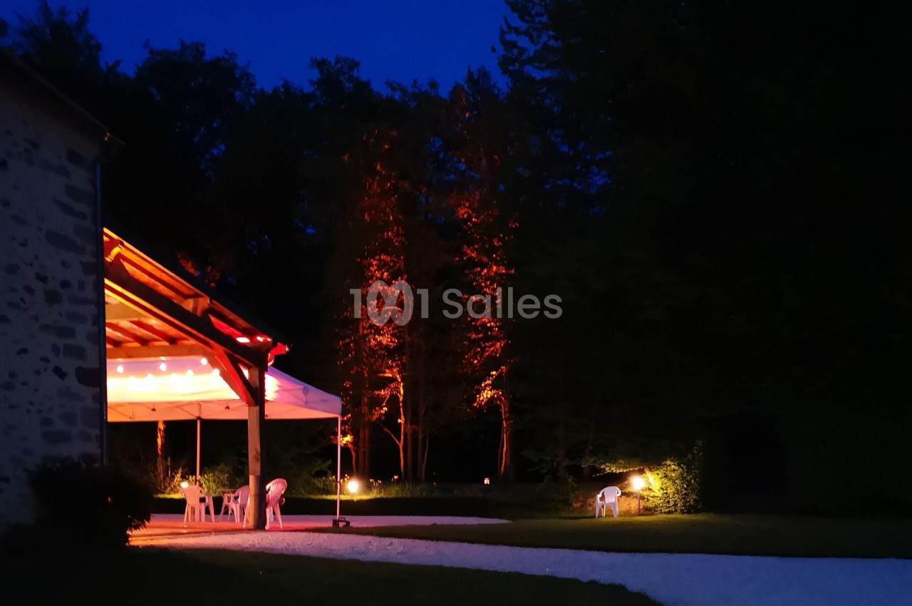 Terrasse éclairée sous un auvent en soirée, entourée d'arbres illuminés et d'un chemin en gravier.