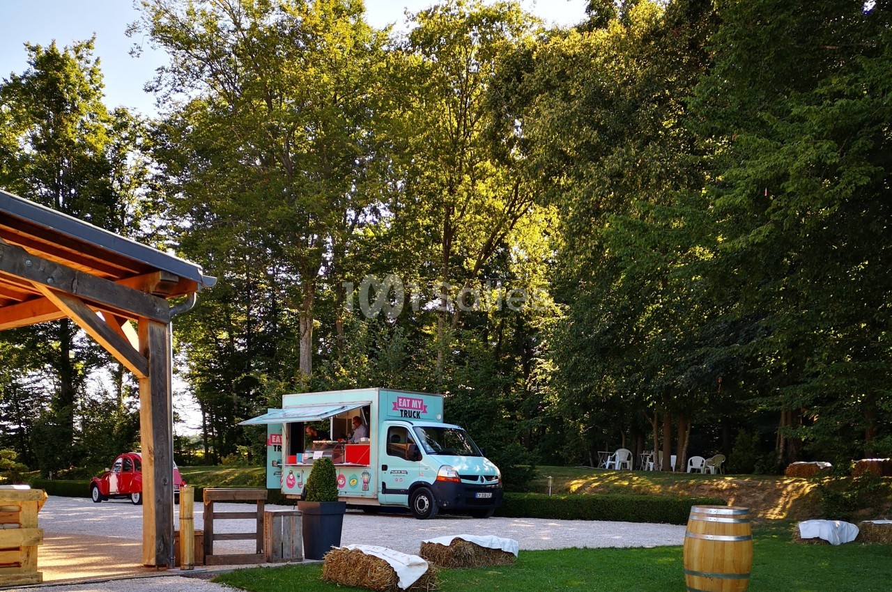 Camion de restauration coloré stationné dans un espace verdoyant près d'une terrasse en bois et de tables extérieures.