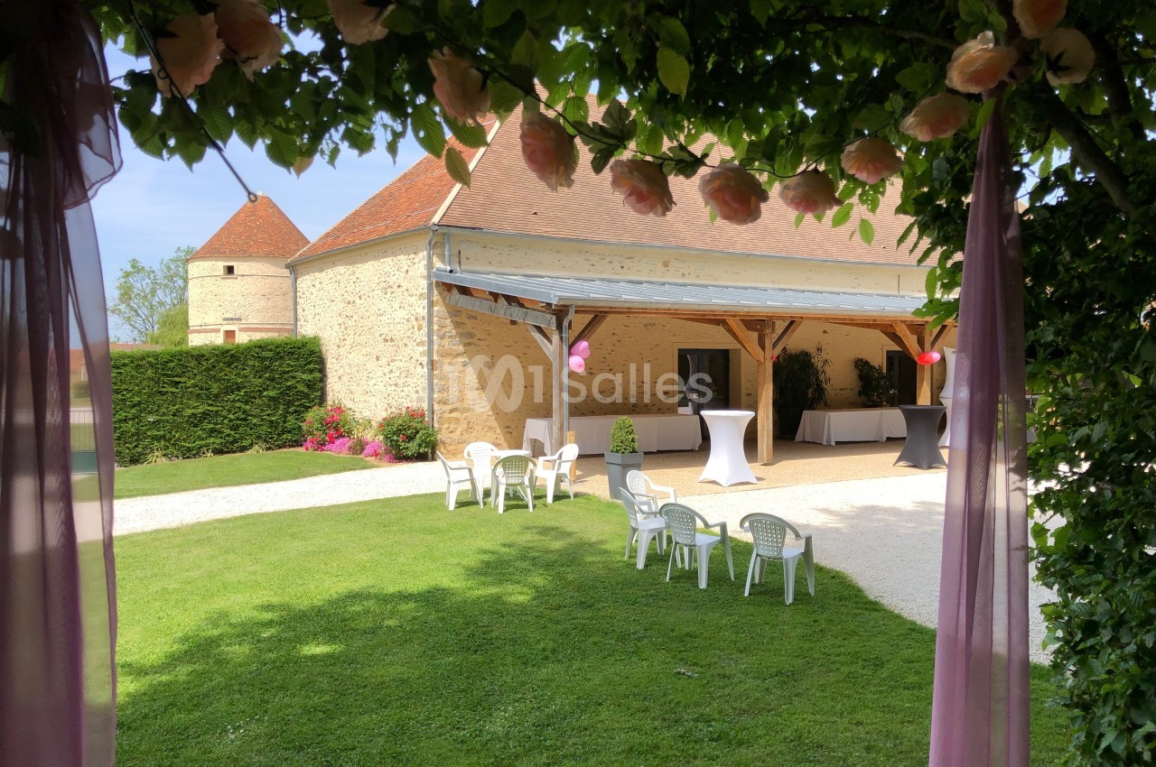 Vue d'une cour verdoyante avec une grange en pierre, des chaises blanches et des tables hautes décorées.