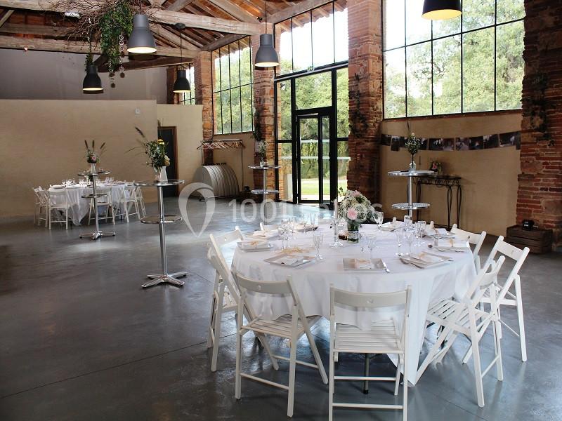 Salle de réception lumineuse avec tables rondes dressées, chaises blanches et grandes baies vitrées donnant sur un extérieur…