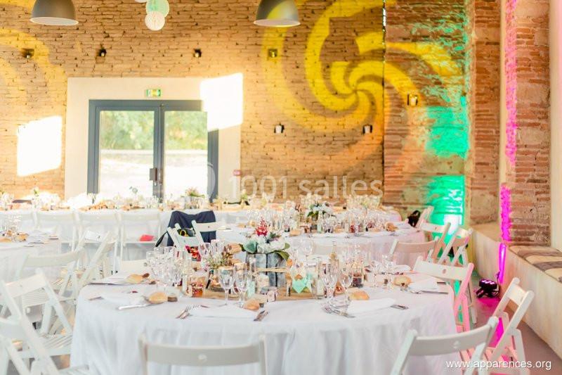 Salle de réception décorée avec des tables rondes dressées, chaises blanches et éclairages colorés sur les murs en briques.