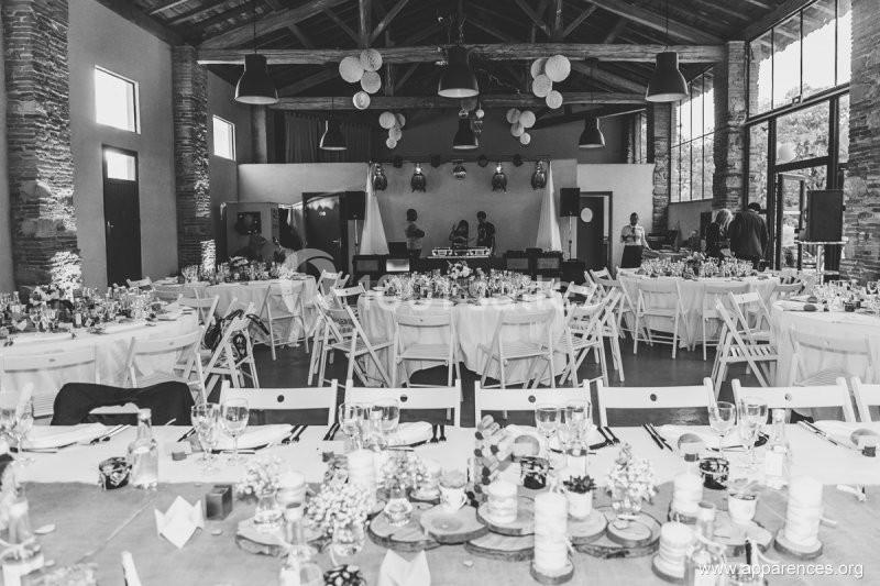 Salle de réception décorée avec des tables rondes dressées, chaises blanches et une scène pour les musiciens.