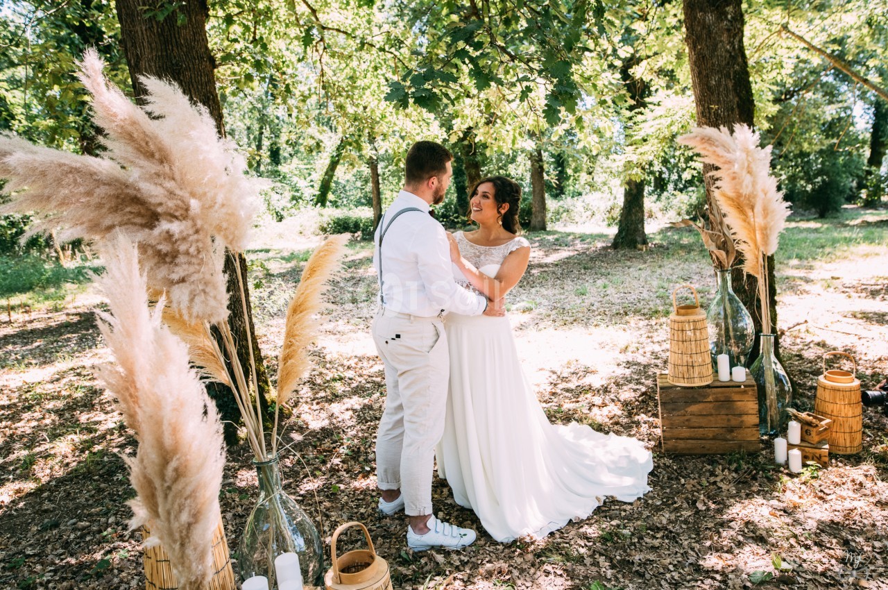 Un couple en tenue de mariage se tient sous des arbres, entouré de décorations naturelles et de pampas.