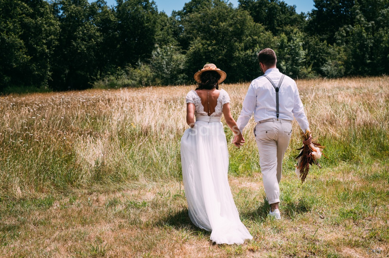 Un couple en tenue de mariage marche main dans la main dans un champ ensoleillé bordé d'arbres.