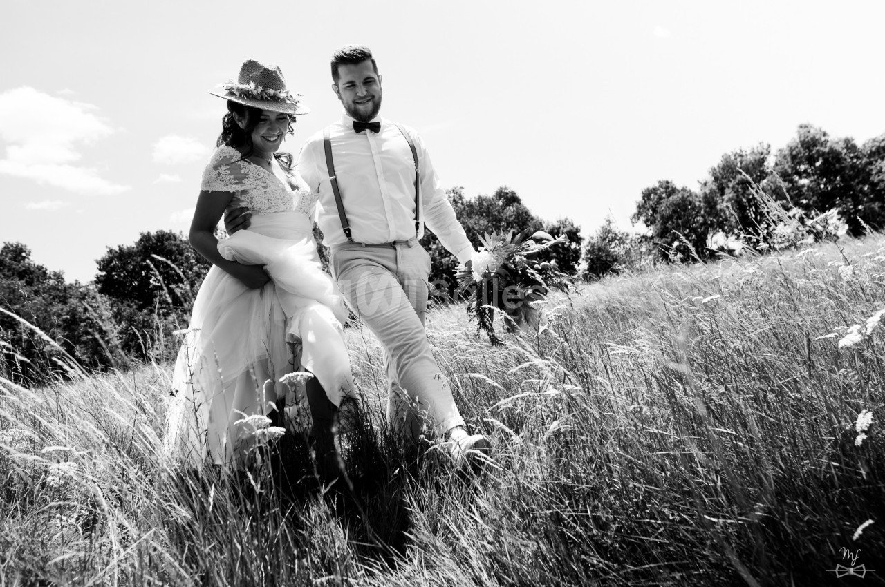 Un couple en tenue de mariage marche dans un champ herbeux sous un ciel dégagé.