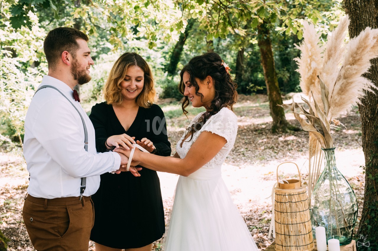 Un couple échange des rubans lors d'une cérémonie en plein air, accompagné d'une officiant sous des arbres.