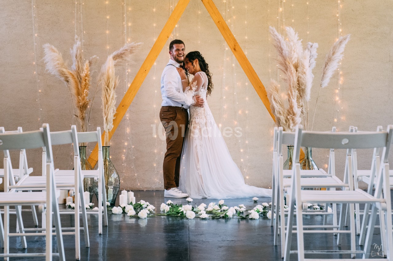Un couple de mariés pose devant une arche en bois décorée de pampas et de lumières, entouré de chaises blanches.