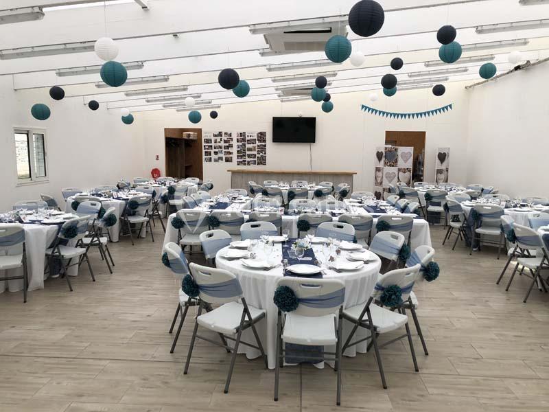 Salle de réception décorée avec des tables rondes dressées, des chaises blanches et des lanternes suspendues.