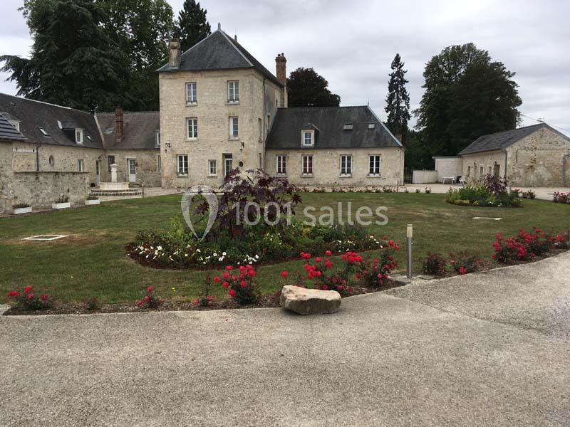 Cour d’un manoir en pierre entourée de parterres fleuris, avec un ciel nuageux et des arbres en arrière-plan.
