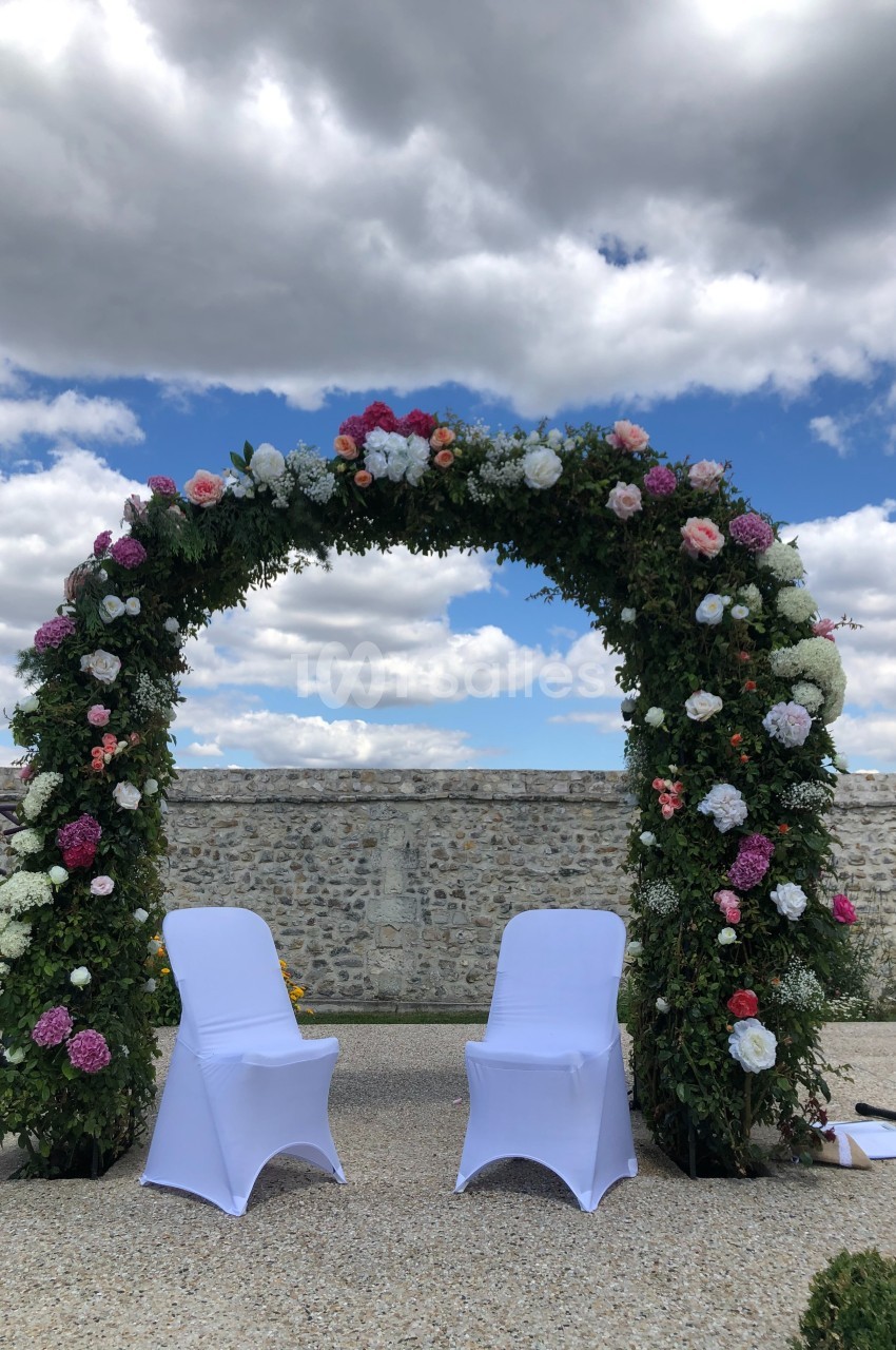 Arche fleurie ornée de roses et pivoines avec deux chaises blanches, installée en extérieur sous un ciel nuageux.