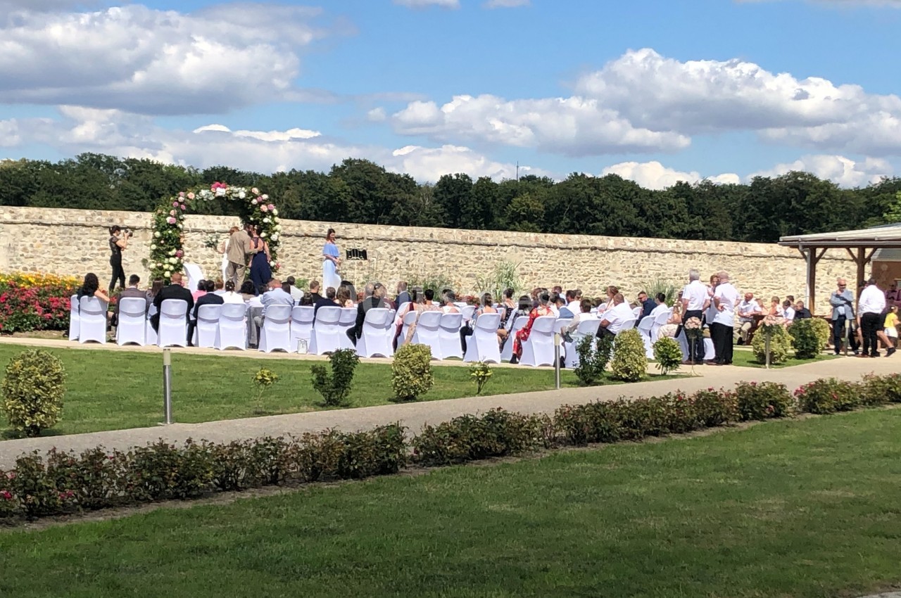 Cérémonie en plein air avec des invités assis devant une arche fleurie, dans un jardin entouré de verdure.