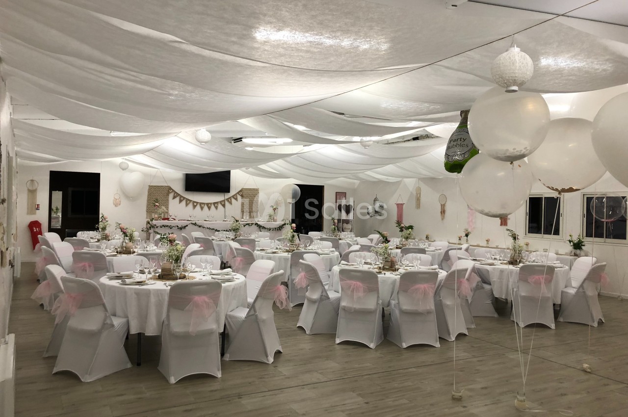 Salle de réception décorée avec des tables rondes, nappes blanches, chaises couvertes et ballons suspendus.