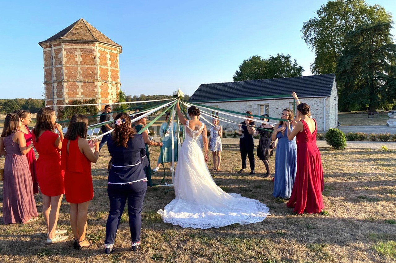 Un groupe de personnes en cercle tient des rubans autour d'une mariée, dans un jardin près de bâtiments anciens.