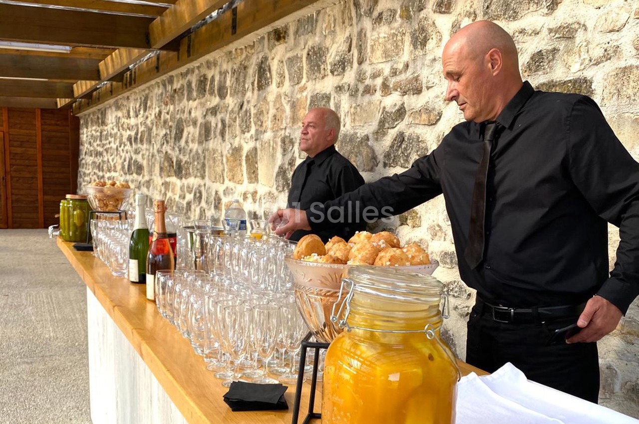 Deux serveurs en tenue noire préparent une table garnie de boissons, verrerie et amuse-bouches devant un mur en pierre.