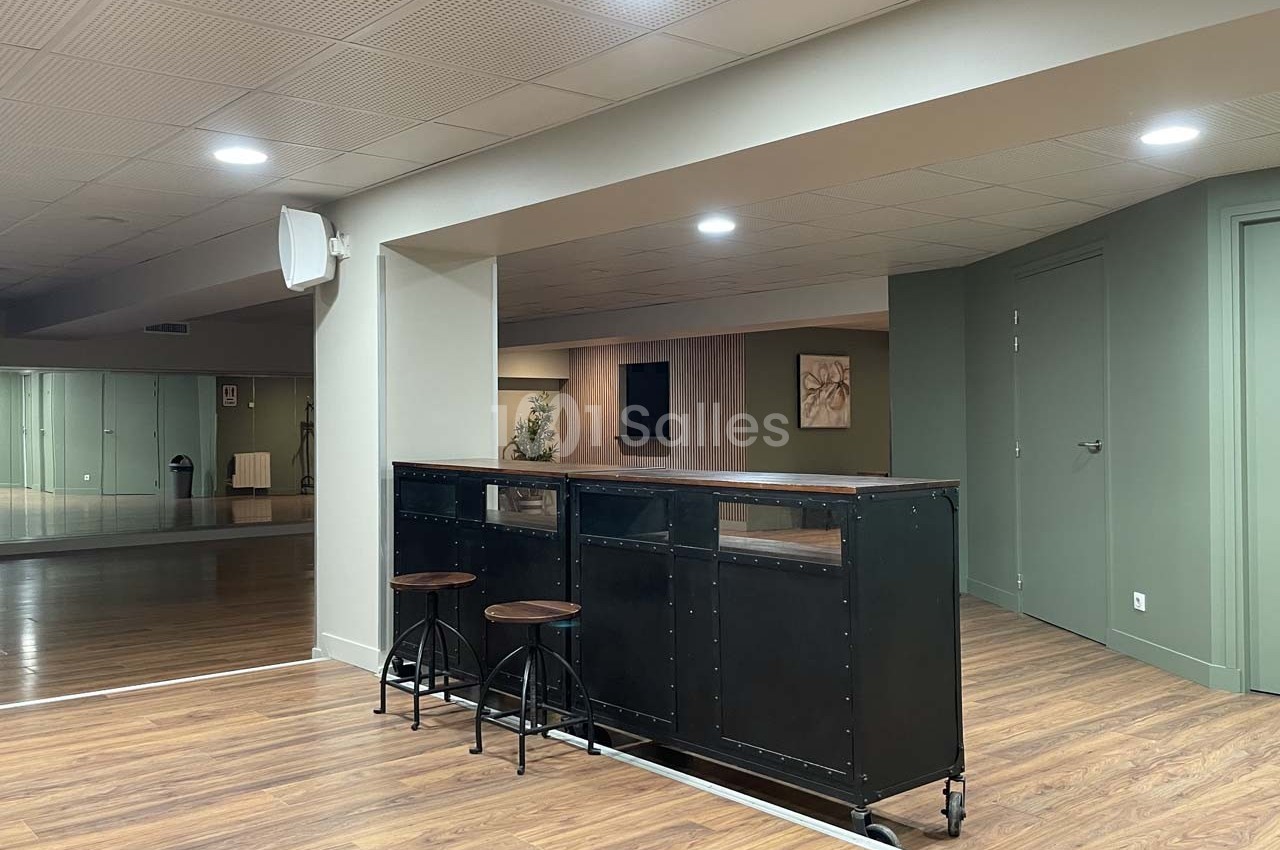 Espace intérieur avec un comptoir noir, deux tabourets hauts, sol en bois et murs verts dans une ambiance lumineuse.