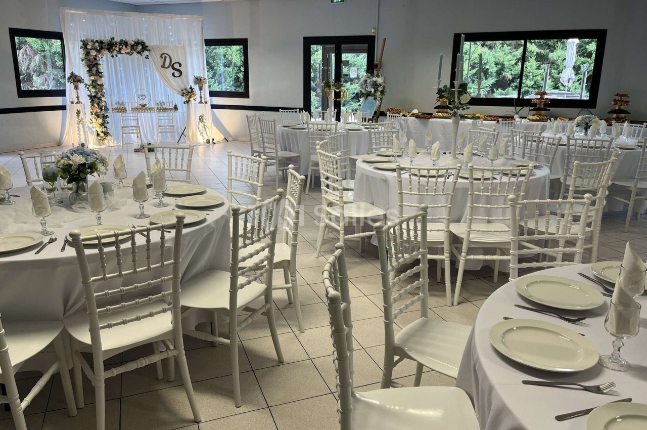 Salle de réception décorée avec des tables rondes dressées, des chaises blanches et un fond fleuri illuminé.