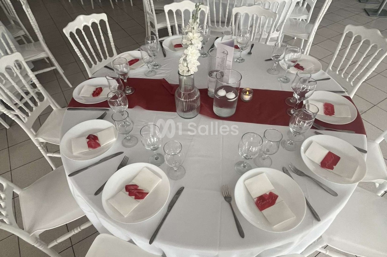 Table ronde dressée avec nappes blanches, chemin de table rouge, vaisselle, verres et pliages de serviettes rouges.