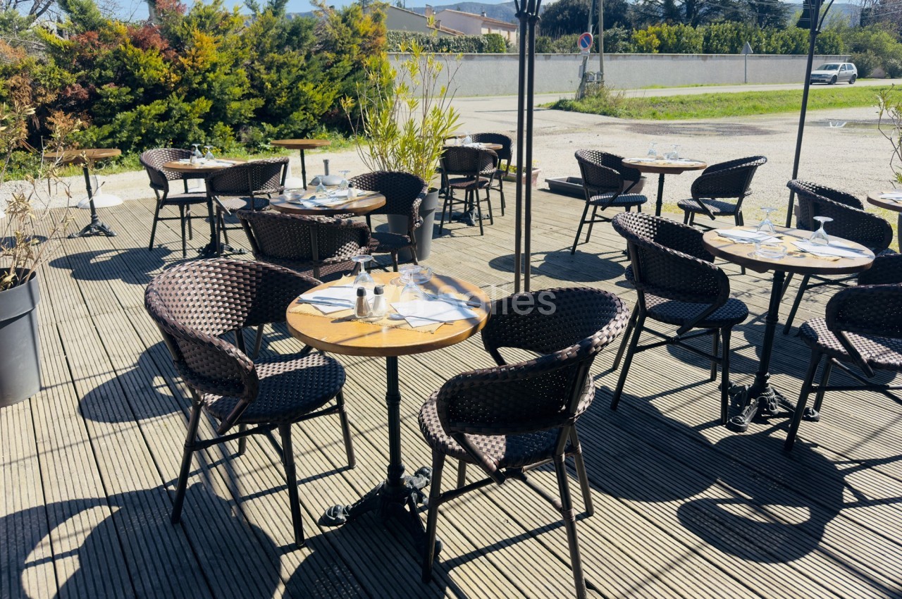 Terrasse ensoleillée avec tables rondes en bois, chaises en osier et couverts disposés, entourée de verdure.