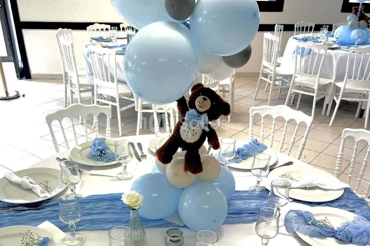 Centre de table avec ballons bleus et blancs, ourson en peluche, nappes bleues et vaisselle disposée sur une table.