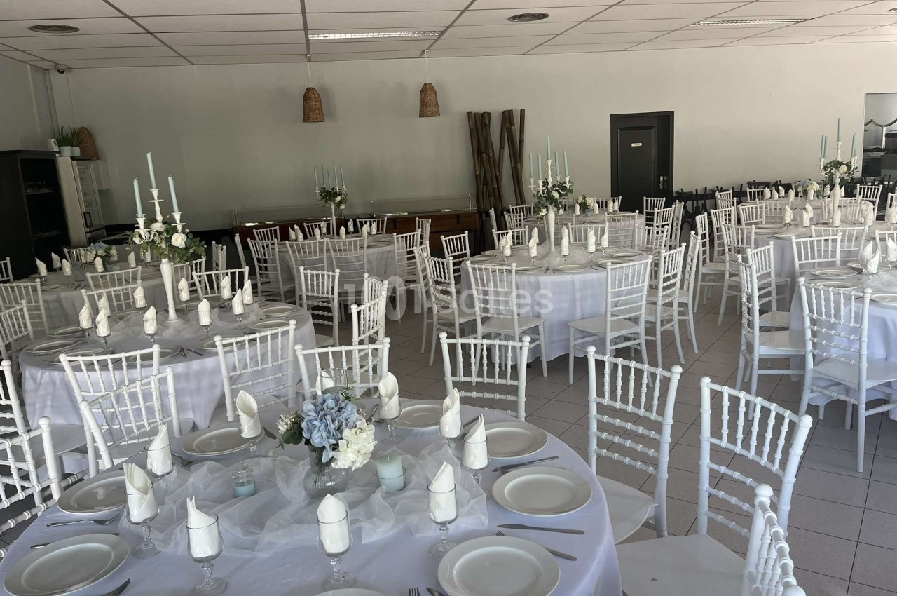 Salle de réception décorée avec des tables rondes dressées, nappes blanches, chaises blanches et centres de table floraux.