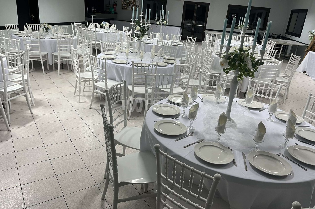 Salle de réception avec des tables rondes dressées, nappes blanches, chaises blanches et centres de table décoratifs.