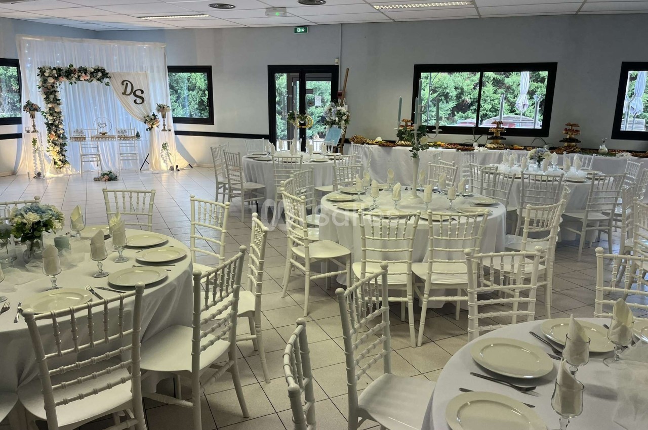 Salle de réception décorée pour un mariage, avec des tables rondes dressées et une arche florale en arrière-plan.