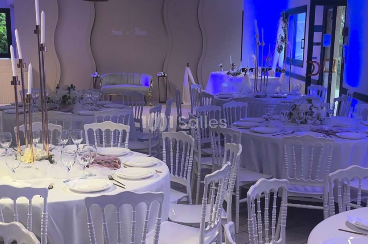 Salle de réception décorée avec des tables rondes dressées, chaises blanches et éclairage bleu en arrière-plan.