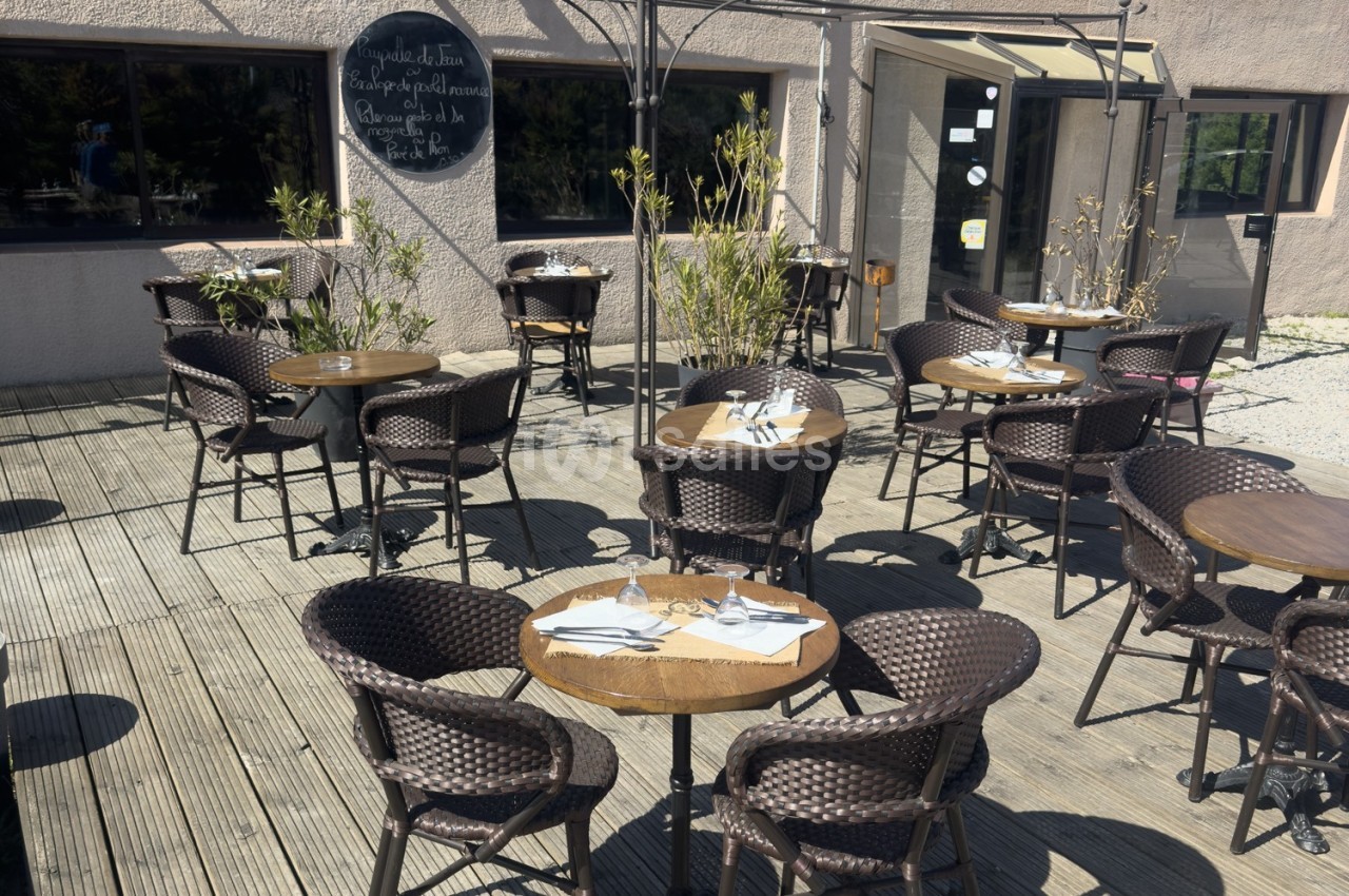 Terrasse en bois avec tables rondes, chaises en rotin et couverts disposés, devant un bâtiment beige.