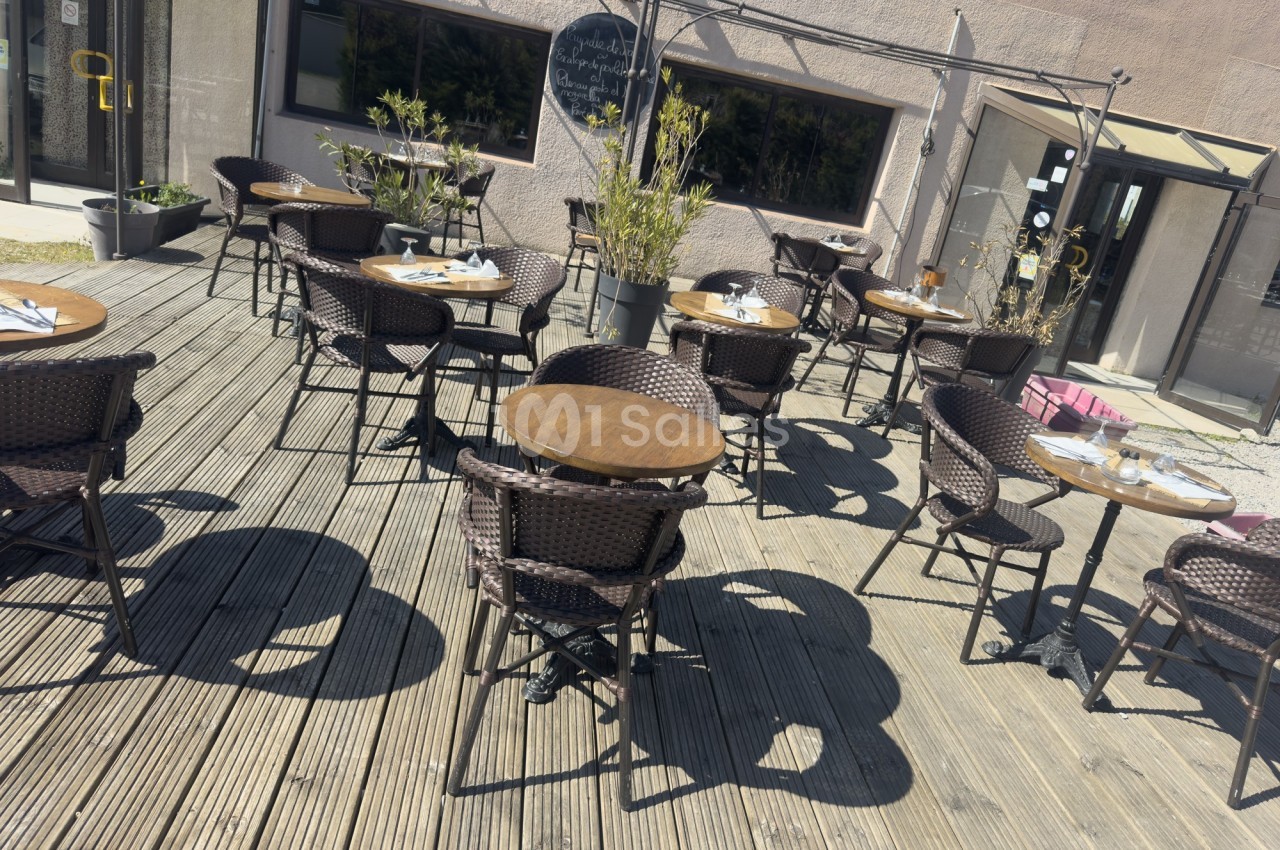Terrasse en bois avec des tables rondes et des chaises en osier, partiellement ombragée par des plantes et des parasols.