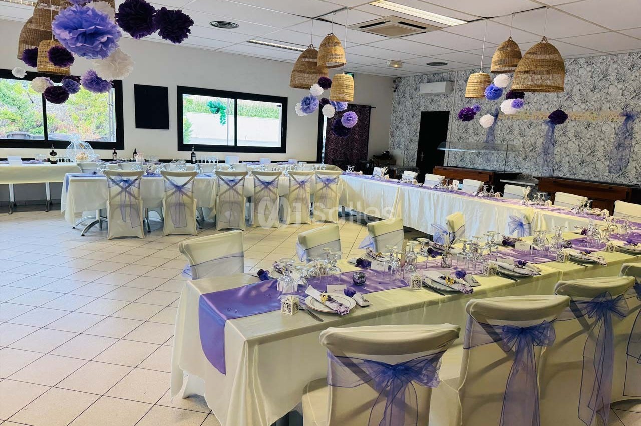 Salle de réception décorée avec des tables dressées, nappes blanches et accents violets, prête pour un événement.